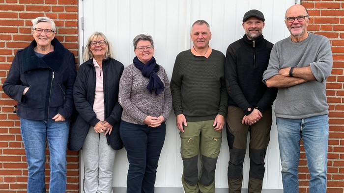 En gruppe af seks personer står sammen ved en mur af røde teglsten.