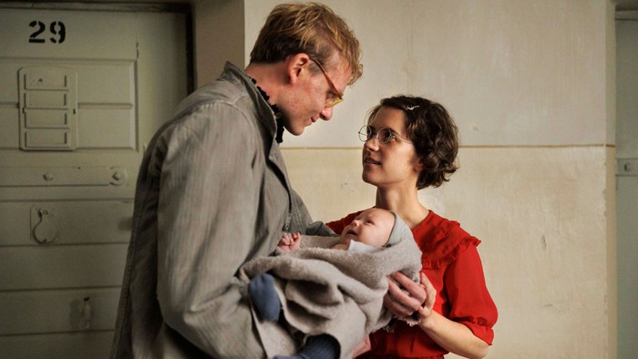 Auf dem Foto sind ein Mann und eine Frau zu sehen, die zwischen sich ein Baby halten. Der Mann (in grauer Jacke) schaut freundlich auf das Kind. Die Frau (im roten Kleid) lächelt den Mann an. Die Szene findet auf einem Gefängnisflur statt.