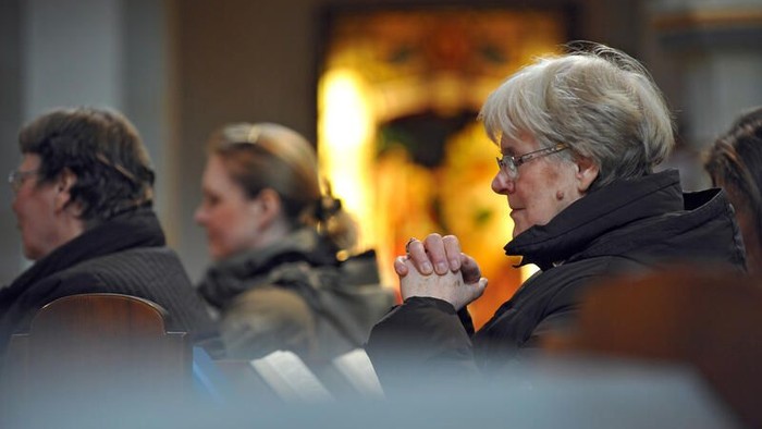 Eine Gruppe von Menschen sitzt in einem Raum, wobei eine Frau deutlich im Vordergrund zu sehen ist.