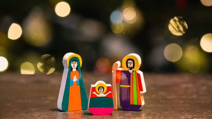 Three colorful wooden figures, likely representing religious or festive characters, are arranged in a row on a surface with blurred lights in the background.