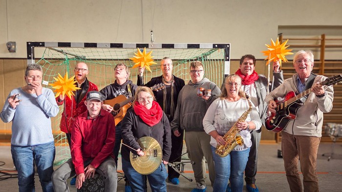 Mehrere Menschen mit Musikinstrumenten spielen in einer Turnhalle vor einem Tor. Dahinter stehen zwei Männer und eine Frau, die große gelbe Sterne hochhalten.