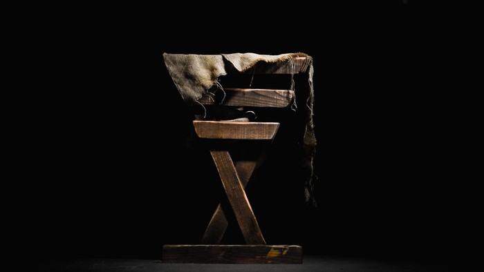 A wooden chair with a blanket draped over the back, set against a dark background.