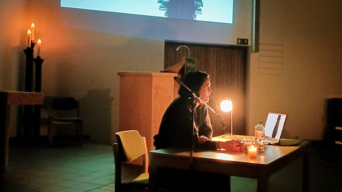 Eine junge Frau sitzt an einem Schreibtisch in einer abgedunkelten Kirche. Im Hintergrund Kerzen und eine Leinwand.