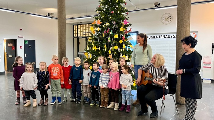 Kinder versammeln sich um einen Weihnachtsbaum mit zwei Frauen, von denen eine sitzt und die andere steht.