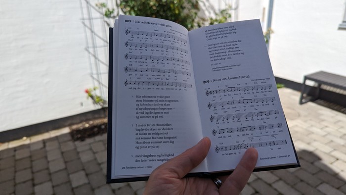 En hånd holder en åben bog med noter og tekst ved en strand