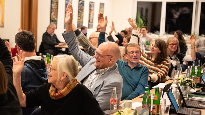 Eine Gruppe von Menschen sitzt an einem Tisch und hebt die Hände, offenbar bei einer Feier oder einem Treffen.