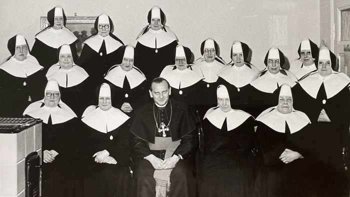 Ein Priester sitzt umgeben von Nonnen in einer Reihe