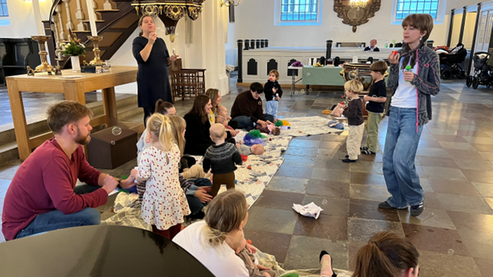 En gruppe mennesker, der sidder og står i et rum med en krystallysekrone og store vinduer.