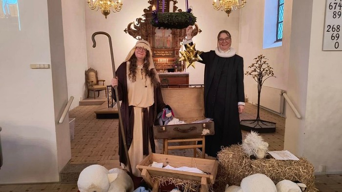 To kvinder, den ene i hyrdekostume og den anden i præstekjole, stående i en kirke i julekrybbe-tableau med krybbe, halmballe, tøjdyr og en adventskrans i baggrunden 