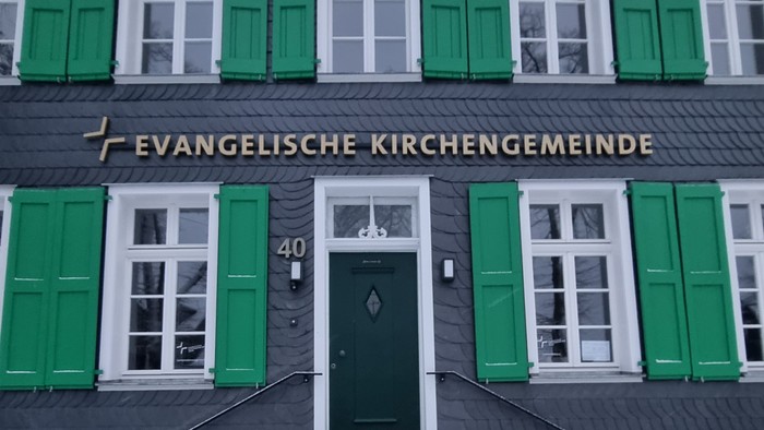 Das Bild zeigt ein Gebäude mit grünen Fensterläden und einer Treppe zum Eingang.