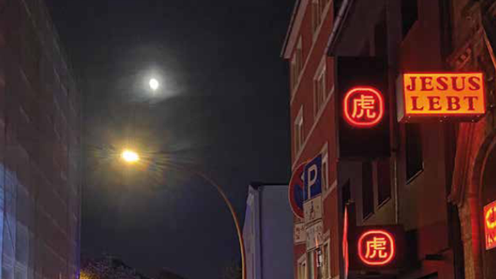 Zeitschriftencover mit einer nächtlichen Straßenszene, auf der Menschen und beleuchtete Schilder zu sehen sind, darunter ein auffälliges Schild mit der Aufschrift *„Jesus liebt dich“*.