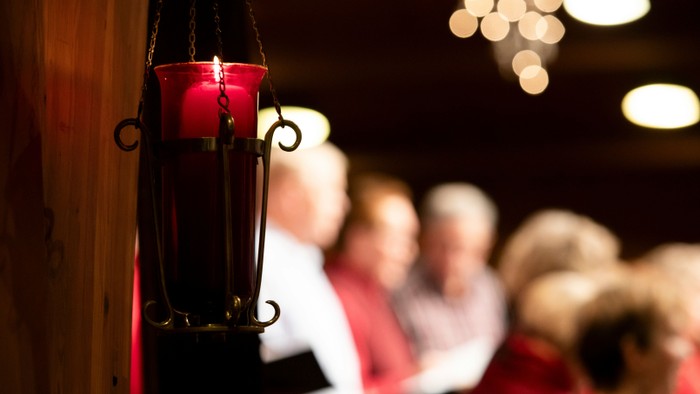 Eine rote Kerze in einer Laterne steht im Vordergrund, während im Hintergrund eine Gruppe von Menschen zu sehen ist.