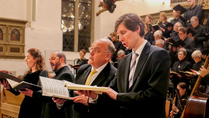 Eine Sängerin und drei Sänger in einer Kirche. Im Hintergrund ein gemischter Chor in Schwarz