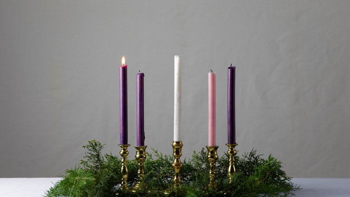 Four candles in brass holders, three unlit and one lit, surrounded by greenery.