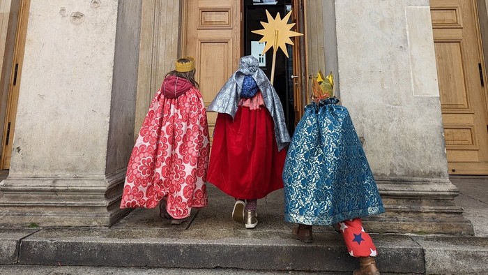 Drei Kinder in farbenfrohen Gewändern sitzen auf Treppenstufen vor einer Tür.