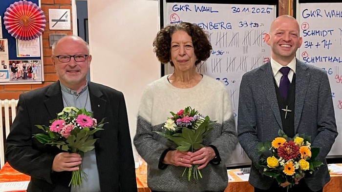 Vier Personen mit Blumensträußen stehen gemeinsam vor einer Gemeinschaftstafel mit Wahlergebnissen.
