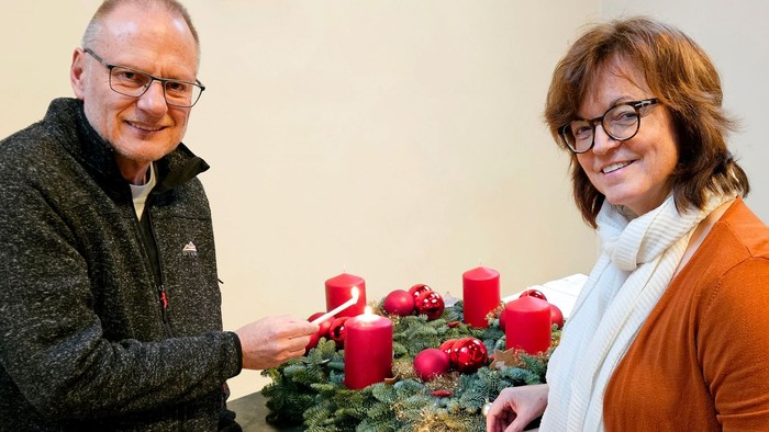 Zwei Menschen stehen an eine Adventskranz, beide entzünden eine Kerze.