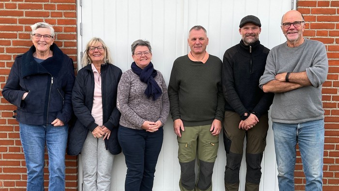 En gruppe af seks personer står sammen ved en mur af røde teglsten.