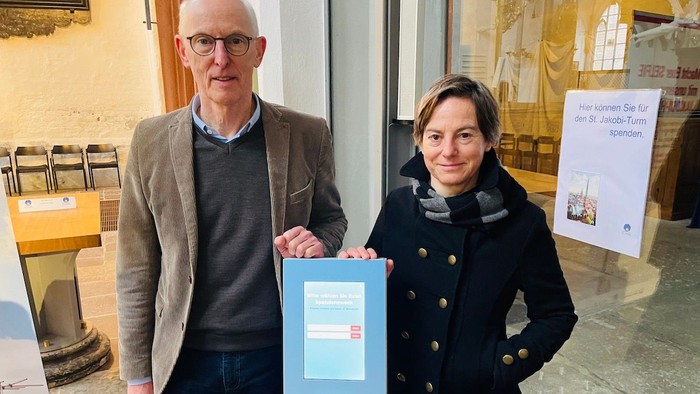 Dr. Jan Verwey, Vorsitzender Stiftung 7Türme+, und Pastorin Bärbel Reichelt, stehen in St. Jakobi an der Spendensäule in der St.-Jakobi-Kirche und lächeln in die Kamera.