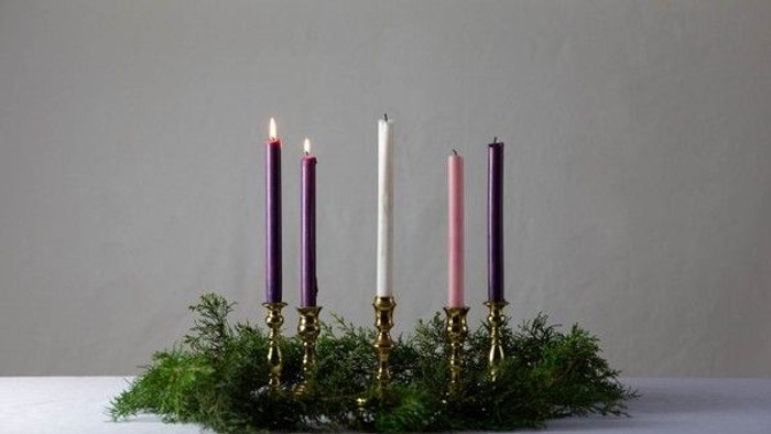 Four lit candles in gold holders on greenery.