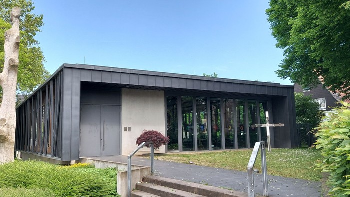 Modernes Gebäude mit großen Glasfenstern und -türen, umgeben von Grünflächen und Bäumen.