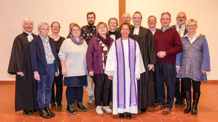 Eine Pfarrerin in heller Amtstracht steht im Zentrum einer gemischten Gruppe, dabei zwei weitere Pfarrerinnen im Talar. Alle lächeln.
