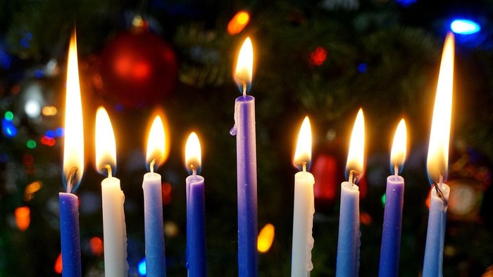 Neun brennende Kerzen in verschiedenen Farben, in einer Reihe angeordnet, mit einem unscharfen Hintergrund aus bunten Lichtern.