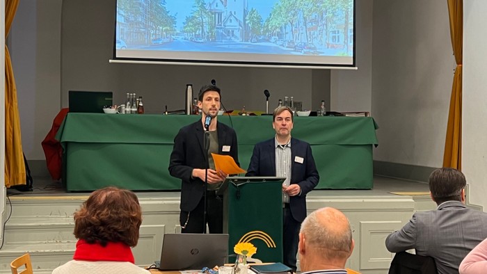 Zwei Männer an Rednerpulten sprechen zu einem Publikum mit einer Präsentationsleinwand hinter ihnen.