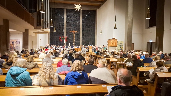 Viele Menschen sitzen in einer Kirche, aus einer hinteren Bank gesehen. Im Altarraum ist ein Posaunenchor klein zu erkennen. Darüber hängt ein Herrnhuter Stern.