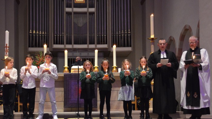 Foto (Abdinghof): Kinder der Attenborough – School (grün) und der Lutherschule (weiß) präsentieren stellvertretend ihre Christingles mit den Pfarrern Nick Adley (r.) und Eckhard Düker
