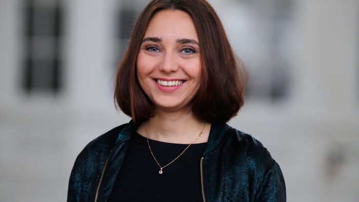 Junge Frau mit kurzen braunen Haaren, die lächelt und eine dunkle Jacke sowie eine Halskette trägt.