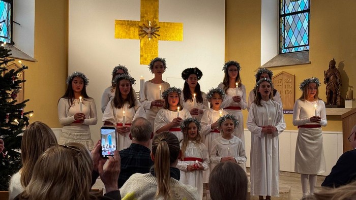 Børn i hvide kjoler og blomsterkranse, der synger i kirken.