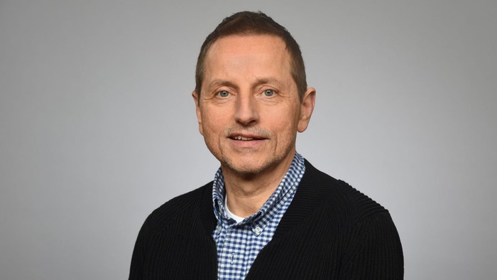 Ein Mann mit dunklem Haar und Bart, der ein schwarzes Jackett und ein blau-weiß kariertes Hemd trägt.