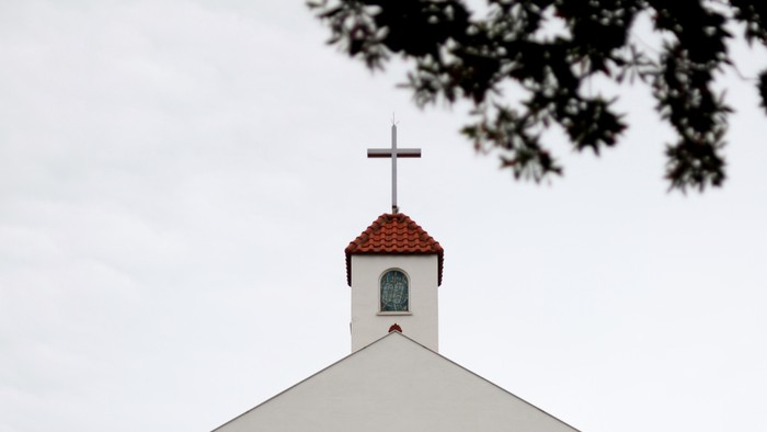 Weißer Kirchturm mit roter Kappe und Kreuz, teilweise von Bäumen verdeckt