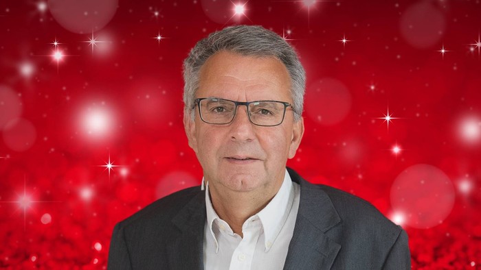 Ein Mann mit Brille und Anzug vor rotem Hintergrund mit weißen Punkten.