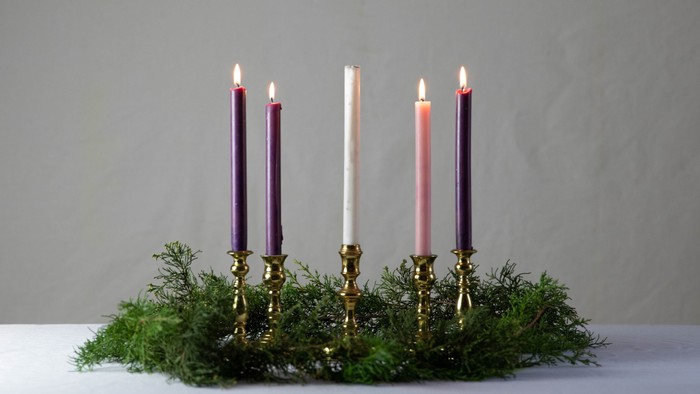 Five lit candles in brass holders surrounded by greenery