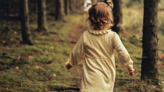 Kind mit lockigem Haar und hellem Kleid läuft auf moosbedecktem Waldweg zwischen Bäumen.