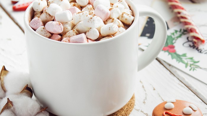Eine festliche Szene mit einer Tasse heißer Schokolade, die mit Marshmallows garniert ist, umgeben von Weihnachtsdekorationen wie einem Lebkuchenmann-Plätzchen, Zuckerstangen und Christbaumkugeln.