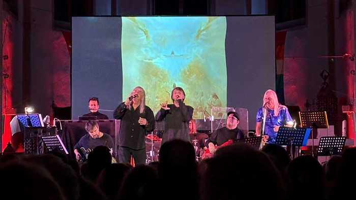 Die Musiker treten auf der Bühne mit einer großen Leinwand hinter sich auf.