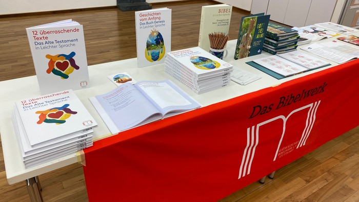 Büchertisch mit einem roten Tuch an der Frontseite, auf dem das Logo des Bibelwerkes weiß zu sehen ist. Darauf die Neuerscheinung von November 2025. Parkettboden. Weiße Wände.