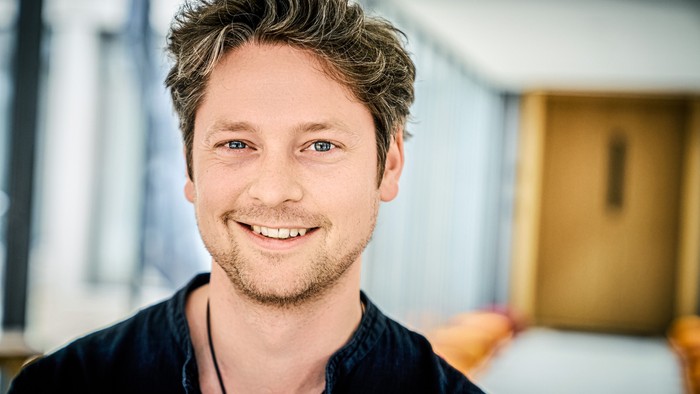 Lächelnder Mann mit lockigem Haar, der drinnen in einer lässigen Umgebung steht.