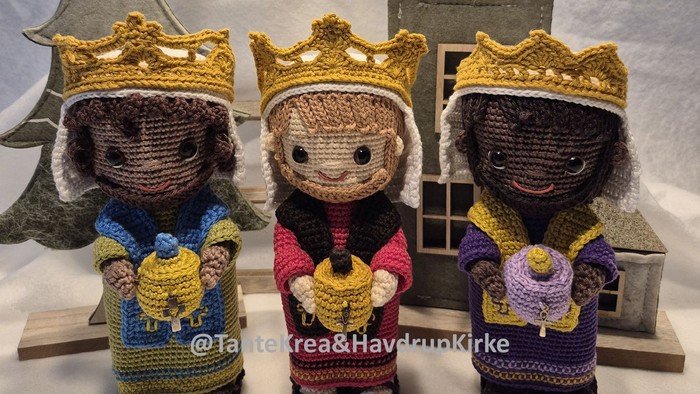 Tre hæklede figurer med kroner, der hver holder en gave, placeret foran et træhus og en juletræ.