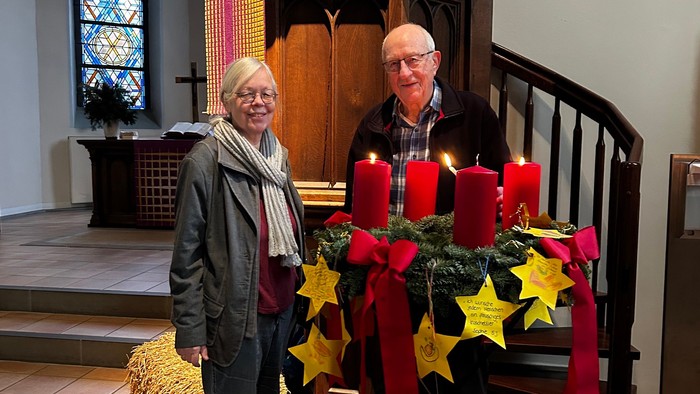 Zwei Personen stehen neben einem geschmückten Adventskranz und einer Kanzel in einer Kirche während der Weihnachtszeit.