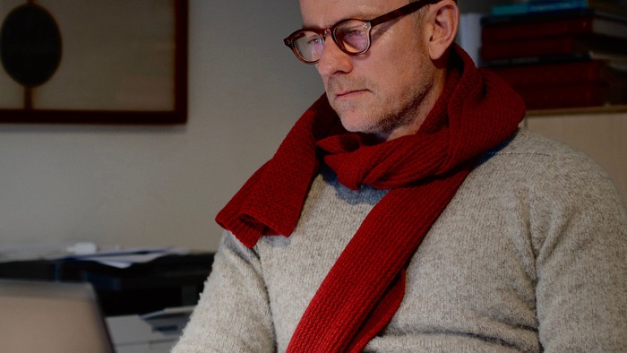 Mand med briller og rød halstørklæde, siddende ved et skrivebord med en computer.