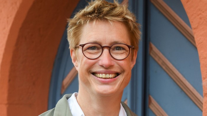 Eine fröhliche Person mit Brille, einem grünen Mantel und einem weißen Oberteil vor farbenfrohen architektonischen Bögen.