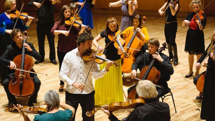 Mitglieder eines Streichorchesters, die gemeinsam in einem Konzertsaal musizieren und sich auf ihre Instrumente konzentrieren.