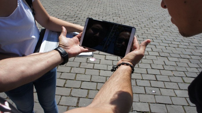 Menschen blicken auf ein Smartphone, dass ein Mensch weit von sich hält, so dass alle auf das Display schauen können. Gepflasterter Untergrund.