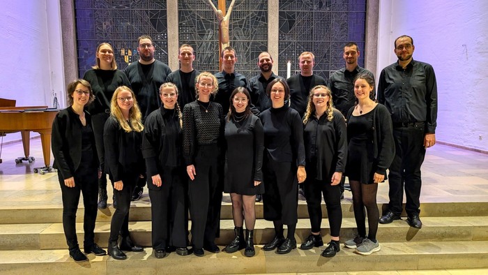 Ein gemischter Chor mit neun jungen Frauen und sieben jungen Männern steht auf zwei Treppenstufen vor einem modern gestalteten Altarraum. Alle tragen schwarz.