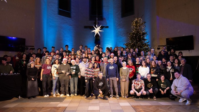 Eine große Gruppe posiert fröhlich für ein festliches Weihnachtsfoto mit einem geschmückten Weihnachtsbaum und einem Sternen-Hintergrund.