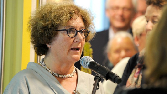 Eine Frau mit Brille spricht in ein Mikrofon bei einer öffentlichen Redeveranstaltung, umgeben von einem Publikum.